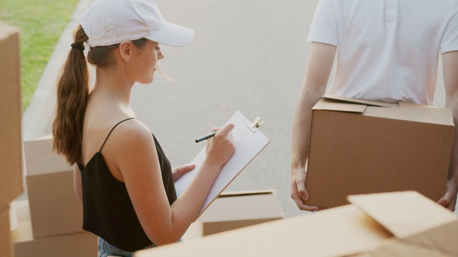 A young woman wearing a white baseball cap and black sleeveless top is inside a property, taking notes on a clipboard with a pen, likely during a home relocation or packing process. In front of her, an unidentified person, dressed in a white T-shirt, is holding a medium-sized cardboard box, which is secured with packing tape. Around them, there are several other cardboard boxes of varying sizes, some stacked, some on the floor, indicating an ongoing packing and moving operation. The environment appears well-lit with natural light, and the scene is set on a paved or concrete surface just outside or near the entrance of a building. Various packing materials, such as cardboard boxes and possibly bubble wrap or plastic, are visible, suggesting careful preparation for furniture transport and home relocation. This image illustrates the process of packing, organizing, and planning involved in professional removals, as performed by Man and Van Bethnal Green, supporting efficient loading and transportation for house moves.