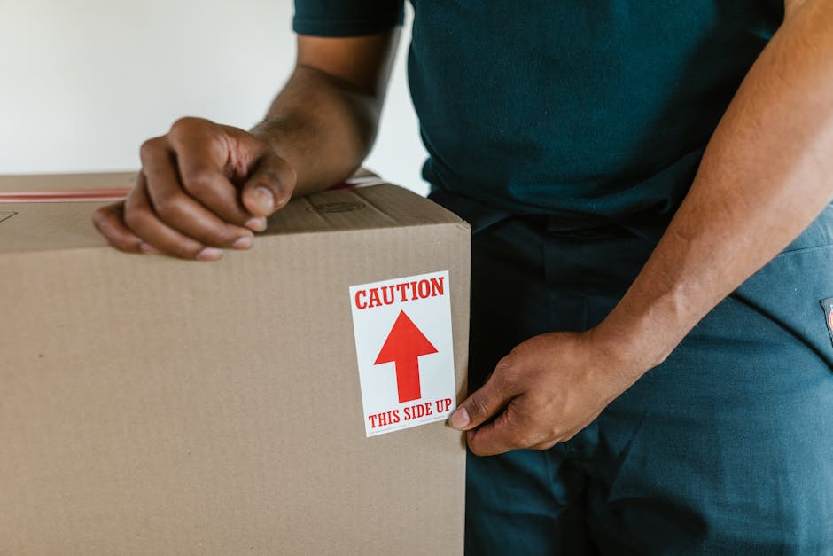 A person wearing a dark green T-shirt and dark trousers is handling a cardboard moving box indoors, near a doorway or entryway. The individual is using both hands to hold the box, which is sealed and reinforced with packing tape, indicating careful packing for a home relocation. A prominent red and white label on the box reads 'CAUTION THIS SIDE UP' with an upward-pointing arrow, highlighting the importance of upright handling during furniture transport and packing and moving processes. The scene suggests a loading or unpacking phase, possibly part of a house removal or furniture transport operation coordinated by Man and Van Bethnal Green, who specialize in removals and moving services. The environment features minimal background detail, emphasizing the focus on the handling of the packed box, with the setting implying an organized interior space prepared for a household move.