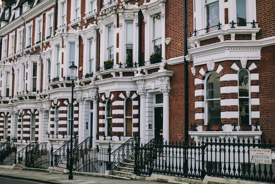 The image depicts a row of Victorian-style terraced houses with red brick facades and elaborate white decorative trim around the windows and doors, typical of homes in Bethnal Green. Each residence features tall, narrow arched windows with white frames and black wrought iron railings along the small front steps leading to the entrances. A black street lamp is positioned on the pavement in front of the buildings, illuminating the area during nighttime. The ground floor windows have flower boxes with greenery and flowers, adding a touch of color. The sidewalk is paved with concrete slabs, and a street sign indicating 'Hornton Street W8' is visible on the right side, near the railings. The scene is well-lit, with natural daylight highlighting the architectural details, and the overall environment suggests a quiet residential area, possibly during a house removal or transport event organized by Man and Van Bethnal Green when preparing for home relocation or furniture transport.