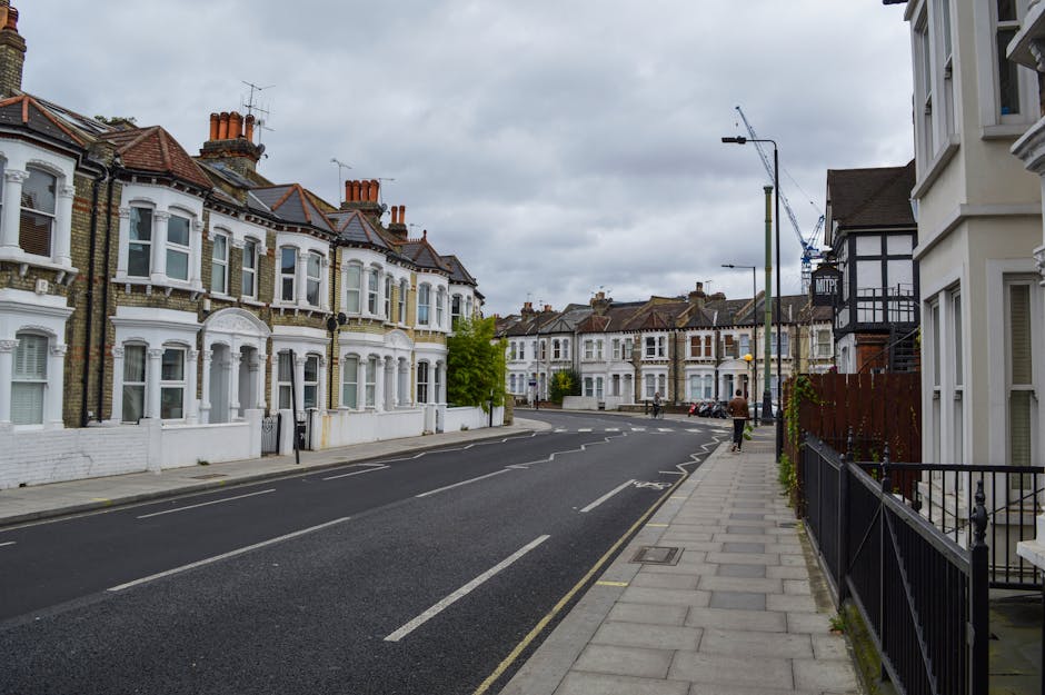 A daytime street view of Victorian terraced houses along Bethnal Green Road featuring white-painted facades, bay windows, and brick chimney stacks, under an overcast sky. The pavement on the right side is lined with a black wrought iron fence and a narrow strip of greenery, with a few pedestrians walking along the sidewalk. The street has a designated lane for vehicles, with painted lines and road markings, and is equipped with street lamps and traffic signs. In the background, there is a construction crane indicating ongoing development nearby. This scene depicts an urban residential area suitable for house removals and furniture transport services, aligning with the context of home relocation and packing and moving processes, as provided by Man and Van Bethnal Green.