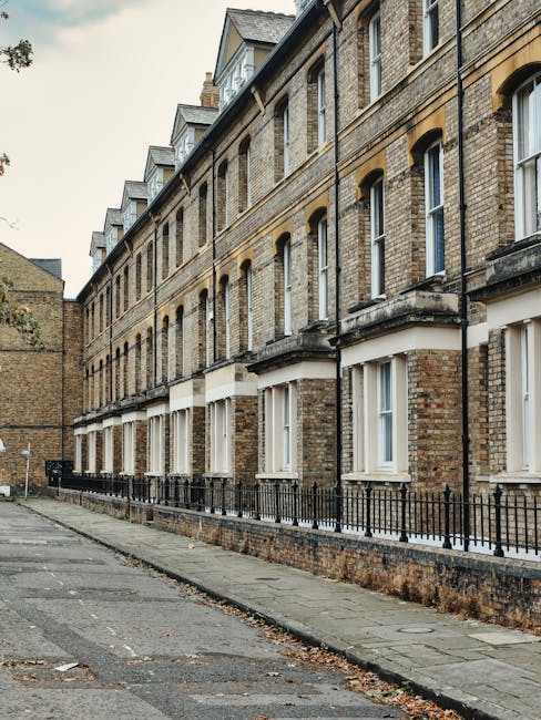 The image depicts a row of Victorian-style terraced houses with red brick facades and elaborate white decorative trim around the windows and doors, typical of homes in Bethnal Green. Each residence features tall, narrow arched windows with white frames and black wrought iron railings along the small front steps leading to the entrances. A black street lamp is positioned on the pavement in front of the buildings, illuminating the area during nighttime. The ground floor windows have flower boxes with greenery and flowers, adding a touch of color. The sidewalk is paved with concrete slabs, and a street sign indicating 'Hornton Street W8' is visible on the right side, near the railings. The scene is well-lit, with natural daylight highlighting the architectural details, and the overall environment suggests a quiet residential area, possibly during a house removal or transport event organized by Man and Van Bethnal Green when preparing for home relocation or furniture transport.
