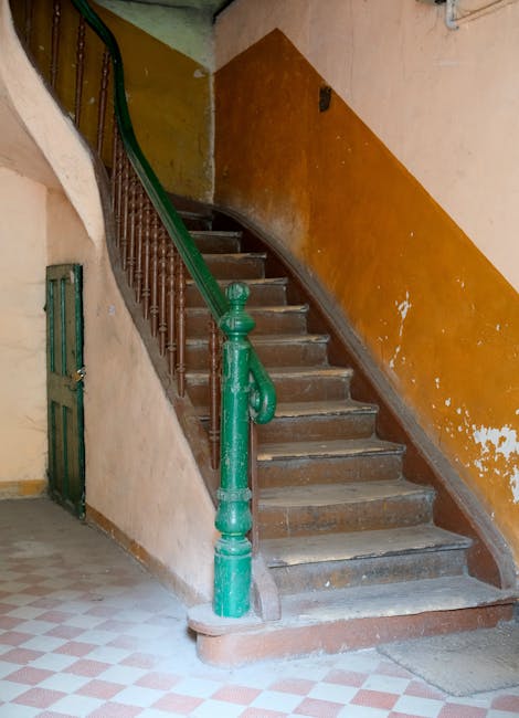 Interior view of a narrow staircase inside a building, featuring aged wooden steps with visible wear and tear, a green metal banister with ornate detailing, and a beige wall with an orange stripe running along the middle. To the left of the staircase, there is a small green metal gate leading to a basement or storage area. The floor at the base of the stairs has a checkered tile pattern in red and white. The lighting appears natural, and the environment suggests an older residential property. During a home relocation or furniture transport, [COMPANY_NAME] would typically handle tasks like navigating tight staircases such as this, ensuring safe loading and moving of household items through narrow, confined spaces, possibly using protective blankets and appropriate lifting techniques.