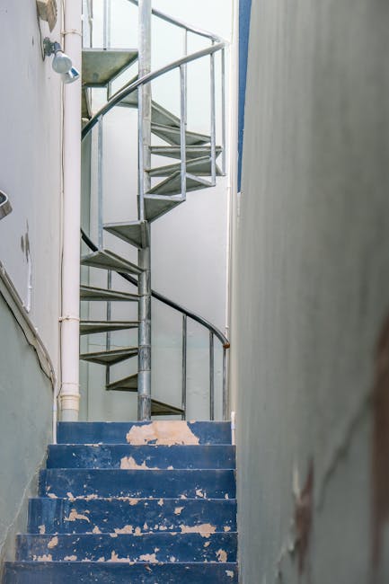 A narrow indoor staircase with blue-painted, worn steps leading up to a small landing where a metal spiral staircase begins. The blue paint on the steps is chipped and peeling, revealing the wooden surface beneath. To the left, white pipes run vertically along the wall, and a small wall-mounted lamp is visible at the top of the staircase. The staircase is enclosed by plain, light-colored walls, with one wall partially visible on the right side of the image. This setting appears to be part of a residential property undergoing a home relocation or moving process, where furniture and boxes may need to be carefully navigated through the tight staircase area. The image is associated with professional removals services, such as those provided by Man and Van Bethnal Green, supporting efficient packing and moving logistics in properties with limited access or space constraints.