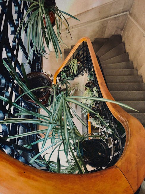 Interior view of a narrow staircase inside a building, featuring aged wooden steps with visible wear and tear, a green metal banister with ornate detailing, and a beige wall with an orange stripe running along the middle. To the left of the staircase, there is a small green metal gate leading to a basement or storage area. The floor at the base of the stairs has a checkered tile pattern in red and white. The lighting appears natural, and the environment suggests an older residential property. During a home relocation or furniture transport, [COMPANY_NAME] would typically handle tasks like navigating tight staircases such as this, ensuring safe loading and moving of household items through narrow, confined spaces, possibly using protective blankets and appropriate lifting techniques.