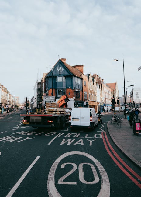 A flatbed delivery truck is parked on a UK urban street, with the rear doors open revealing stacks of wooden pallets and cardboard boxes secured on the truck bed. The truck is positioned at a slight angle across the road, with a white van nearby assisting in loading or unloading items during a home relocation process. Adjacent to the truck, the pavement hosts pedestrians walking, some with shopping bags, and a line of bicycles parked along the curb. The surrounding area features typical town centre buildings, including a prominent blue-stucco corner property with large windows and a peaked roof, and a clear, partly cloudy sky overhead. The street markings show a 20 mph speed limit, and the scene is well-lit with streetlights and natural daylight, reflecting the busy urban environment often associated with house removals and furniture transport logistics, as handled by Man and Van Bethnal Green.