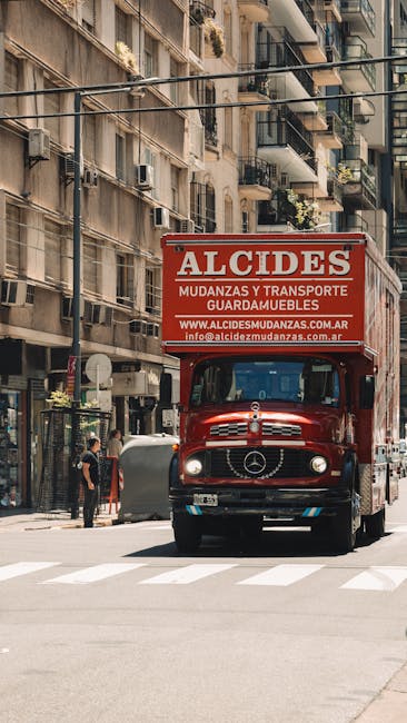 A red moving truck with the branding 'ALCIDES' parked on a city street, ready for a house removal. The truck is positioned close to the curb, with the front facing outward and the rear section aligned parallel to the sidewalk. The truck has a large, rectangular cargo area with signage that includes contact details and a description of moving and furniture transportation services. In the background, multi-storey residential buildings with balconies and small plants visible on some balconies line the street. Pedestrians and parked vehicles are also visible nearby, indicating a busy urban environment. The scene suggests the loading or unloading process during a home relocation, with the truck associated with professional removals by [COMPANY_NAME]. The street is well-lit, and the overall setting captures typical city moving logistics involved in packing, furniture transport, and home relocation tasks.