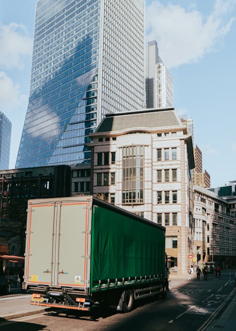 A large green-boxed delivery truck is parked on a city street in front of modern high-rise buildings with glass facades and older mid-rise structures with stone and brick exteriors. The truck is positioned near the curb, with its rear doors closed, ready for loading or unloading. The scene appears to be during the daytime, with natural light illuminating the buildings and vehicle. The street includes pedestrians and other vehicles, and the environment suggests a busy urban setting suitable for a home relocation or furniture transport. The setting aligns with the context of professional removals services offered by Man and Van Bethnal Green, focusing on efficient moving logistics within the city.