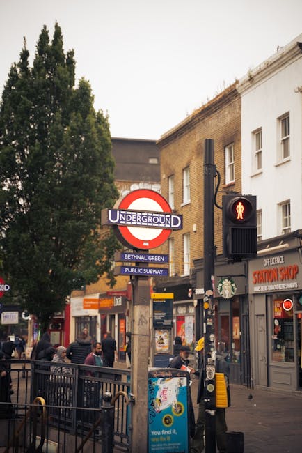 A busy street scene outside a Bethnal Green underground station entrance, featuring a red and blue London Underground roundel sign reading 'UNDERGROUND' with additional signage for 'BETHNAL GREEN' and 'PUBLIC SUBWAY'. In the foreground, a pedestrian crossing signal displays a red standing figure, with a black pole and signal box. The sidewalk is populated with people, some carrying items or waiting to cross, as well as storefronts and commercial signs, including a 'Station Shop' with red signage. To the left, a tall tree with green foliage partially obscures the buildings behind, which are a mix of brick and white facades. The overall environment suggests an urban area with active foot and vehicle traffic, typical of a central London neighbourhood involved in home relocation or furniture transport activities supported by professional removal services like Man and Van Bethnal Green.