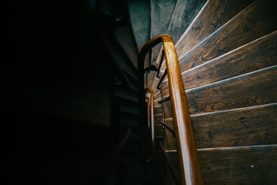 A view from the top of a narrow wooden staircase in a residential property, showing the descending wooden steps with visible grain and finish, and a curved wooden handrail on the right side. The staircase is positioned against a dark wall, with limited natural light illuminating the upper section and creating shadows. The steps lead down to an unseen lower level, and the image captures the confined space typical of interior access points where moving furniture or packed boxes may pose logistical challenges. This setting reflects common access constraints encountered during home relocation or furniture transport, where careful planning is required to navigate tight stairs. Man and Van Bethnal Green specializes in removals, including managing access issues like these during house moves, ensuring safe transportation of belongings through such narrow and difficult areas.