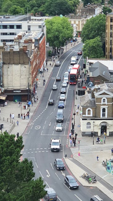 A street scene in Bethnal Green East London featuring a historic clock tower with a rounded top, displaying the words 'East Ham' on a black and white sign. The tower is made of metal with a cylindrica