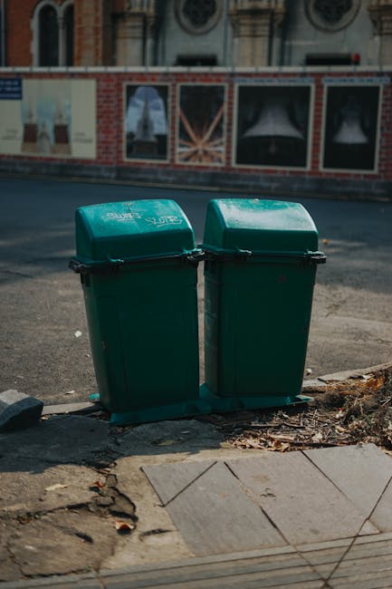 Two green wheeled rubbish bins with black lids positioned on the pavement near the curbside of a residential street, with brick and sash windowed buildings in the background. The bins are secured together with black straps and appear to be used for waste collection. They are situated on a small concrete section adjacent to loose paving stones and dirt, with some debris visible at the base of the bins. The street surface is dark and textured, and the scene is illuminated by daylight, highlighting the details of the waste containers and surrounding environment. This setup suggests a typical urban waste disposal point, relevant to moving and house clearance services provided by Man and Van Bethnal Green, especially in the context of complying with local waste regulations during house removals in Tower Hamlets.