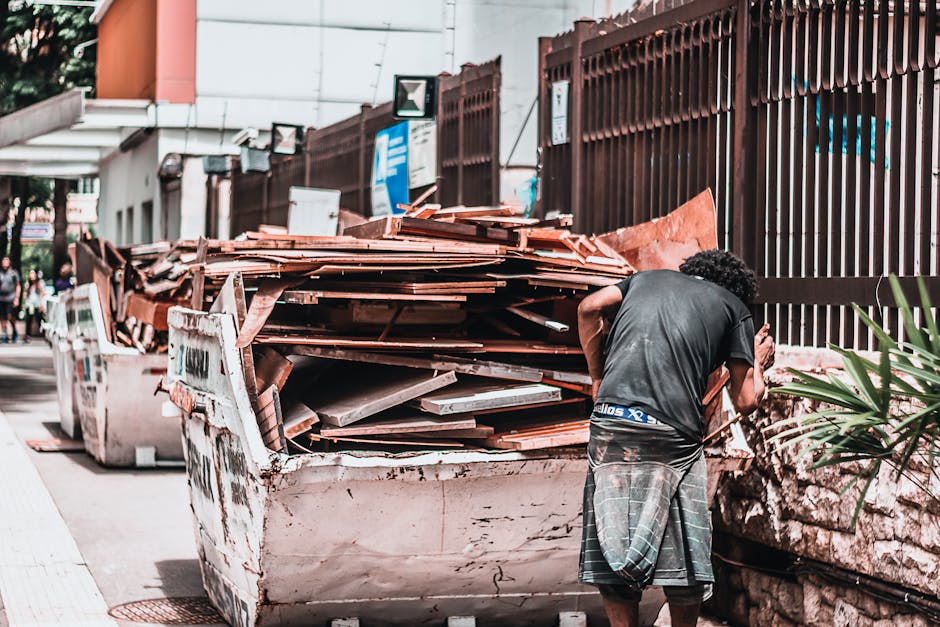 A man with curly black hair, wearing a dark grey T-shirt and checkered shorts, stands on a sidewalk next to a large white skip filled with various building waste materials, including broken wooden planks, tiles, and cardboard packaging. The skip is positioned adjacent to a brown metal fence, with some green plants visible in the foreground. In the background, there are additional skips, a building with signage, and pedestrians walking along the street. The scene appears to be part of a home relocation or clearance process, likely involving packing and disposal of waste during a house move or renovation, relevant to Man and Van Bethnal Green’s removal services. The area is well-lit, with natural daylight illuminating the scene, and equipment such as trolleys or straps are not visible in this specific image.