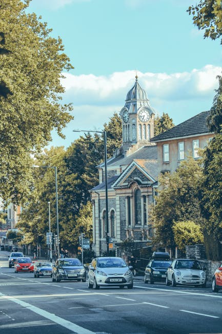 A street scene in Bethnal Green during daylight shows several parked cars along the curb, with a mix of small and medium-sized vehicles including hatchbacks and sedans. In the background, a historic church with a tall clock tower featuring a pointed spire is visible, surrounded by lush green trees and low-rise residential buildings with brick facades and tiled roofs. There is no visible movement or activity involving furniture, boxes, or moving equipment within the scene, as it primarily captures a public road and surrounding architecture. The image appears to be taken during a clear, sunny day, with blue sky and scattered clouds, and natural light illuminating the street and buildings. The scene potentially relates to the context of house removals or furniture transport, with [COMPANY_NAME] offering services in the area, connected to parking permits for home relocations as mentioned in the page title.