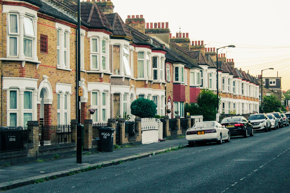 A row of terraced Victorian-style houses in Bethnal Green, Tower Hamlets, with brick facades, bay windows with white trim, and small front gardens enclosed by black metal fences. Several cars, including a white vehicle, are parked along the street pavement adjacent to the houses. Street lamps and utility poles are visible, with overhead wiring crossing the scene. The image depicts a residential area typical for house removals and furniture transport services, with a focus on the exterior environment where loading and logistics activities may take place, supporting the context of home relocation in the Tower Hamlets borough. Man and Van Bethnal Green often operates within such streets for safe and efficient packing, moving, and parking permit management related to house removals.