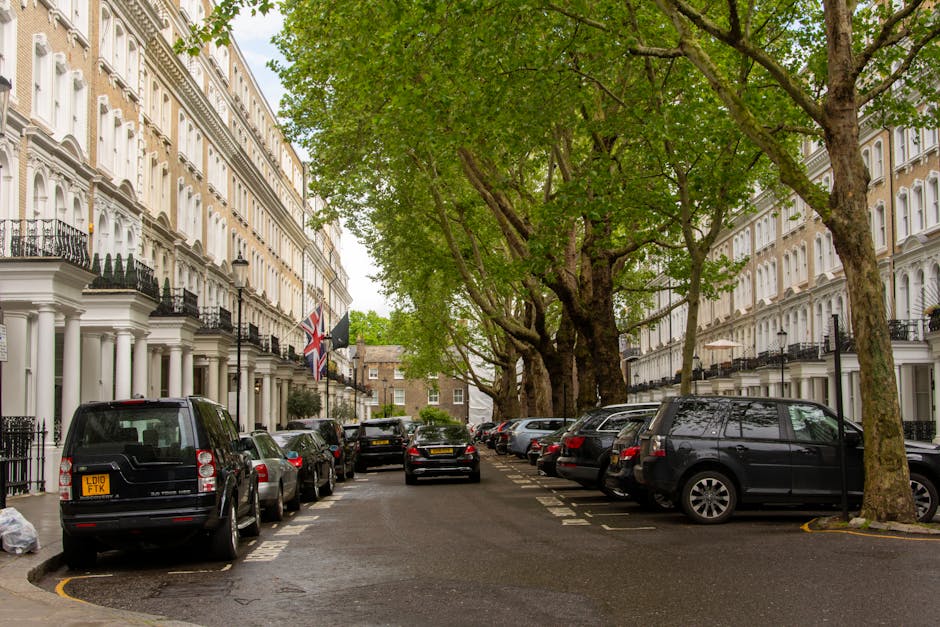 A street scene in Bethnal Green during daylight shows several parked cars along the curb, with a mix of small and medium-sized vehicles including hatchbacks and sedans. In the background, a historic church with a tall clock tower featuring a pointed spire is visible, surrounded by lush green trees and low-rise residential buildings with brick facades and tiled roofs. There is no visible movement or activity involving furniture, boxes, or moving equipment within the scene, as it primarily captures a public road and surrounding architecture. The image appears to be taken during a clear, sunny day, with blue sky and scattered clouds, and natural light illuminating the street and buildings. The scene potentially relates to the context of house removals or furniture transport, with [COMPANY_NAME] offering services in the area, connected to parking permits for home relocations as mentioned in the page title.