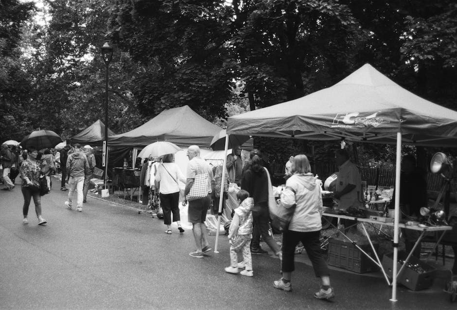 A street scene during a market or event day shows multiple vendor stalls set up under tents with peaked canopies, positioned on a pavement area with a line of large trees and a lamppost in the background. Several people, including adults and children, are walking along the street, some holding umbrellas, suggesting overcast or rainy weather. The vendors' stalls contain various items, possibly including boxes, packaging materials, or small appliances, although specific details are not clearly visible. The scene captures the activity of shoppers browsing and vendors managing their stalls, with some individuals engaged in conversations or examining items. The overall environment consists of outdoor market infrastructure, with tents secured to the ground and a mixed group of pedestrians in casual clothing. The image appears to be part of a broader outdoor event, consistent with community markets or local markets, such as those managed by Man and Van Bethnal Green in the context of home relocation or furniture transport planning.