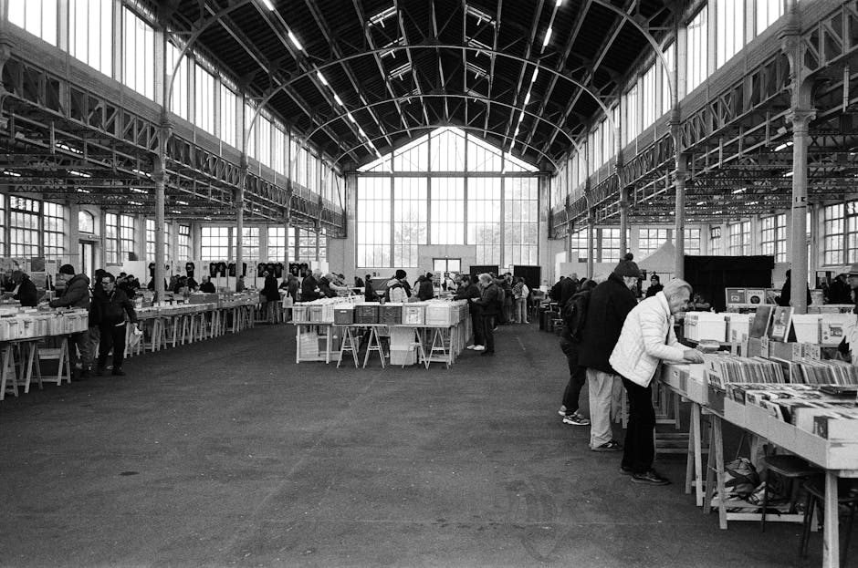 A spacious indoor market hall with a high, arched glass ceiling supported by metal trusses and beams. The interior is well-lit by natural light entering through large windows on the side walls and the front entrance. The floor is covered with a dark, textured surface, and several long tables are arranged in rows, displaying an array of books, packaged in cardboard boxes, plastic crates, and wrapped in protective materials for transport. Customers can be seen browsing and sorting through the books, some lifting boxes or examining items closely. The scene captures a busy atmosphere typical of a market or a book fair, with visitors engaged in selecting items for collection or home relocation. Occasionally, individuals are carrying or loading boxes onto carts or trolleys, indicating a process related to packing and moving. The overall setting emphasizes the logistics involved in home or event-related furniture transport and packing, which is why Man and Van Bethnal Green may provide such removals and moving services.
